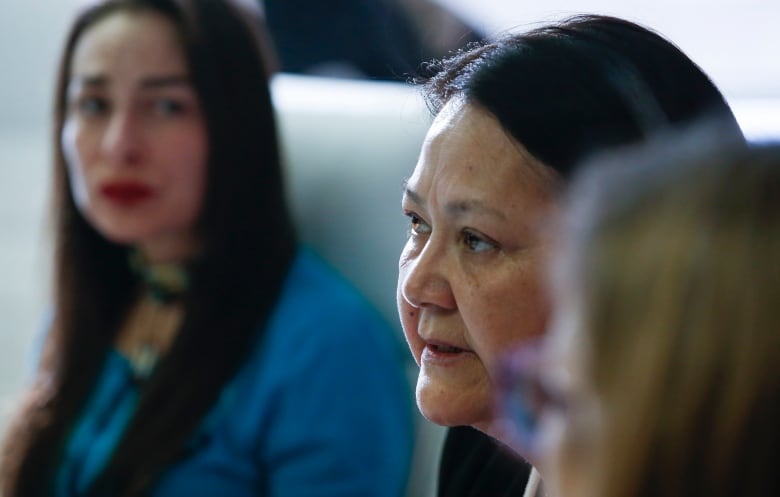 A closeup shows a woman speaking as she sits beside another woman.