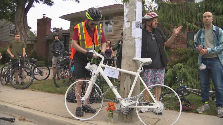 A man chains a white bicycle to a telephone pole on a residential street. Half a dozen cyclists stand around him. It's a grey day