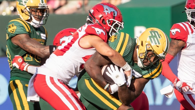 A Black football player in a green-and-yellow uniform cradles a football with both arms, as another player with light skin, wearing a red-and-white uniform, tries to tackle him.