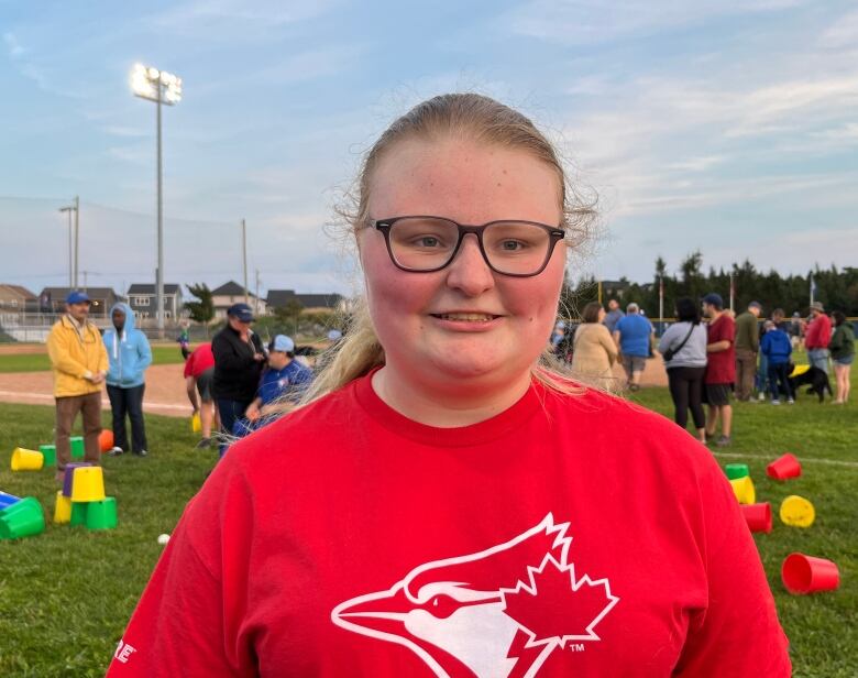 A woman smiles. There are people behind her on a baseball field.