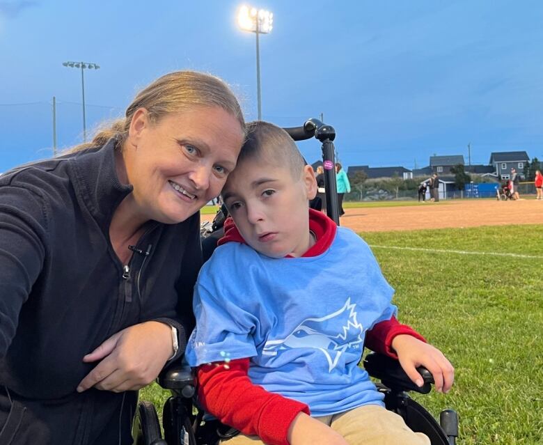 A woman smiles. She kneels down next to a boy who sits in a wheelchair. Behind them is a baseball field.