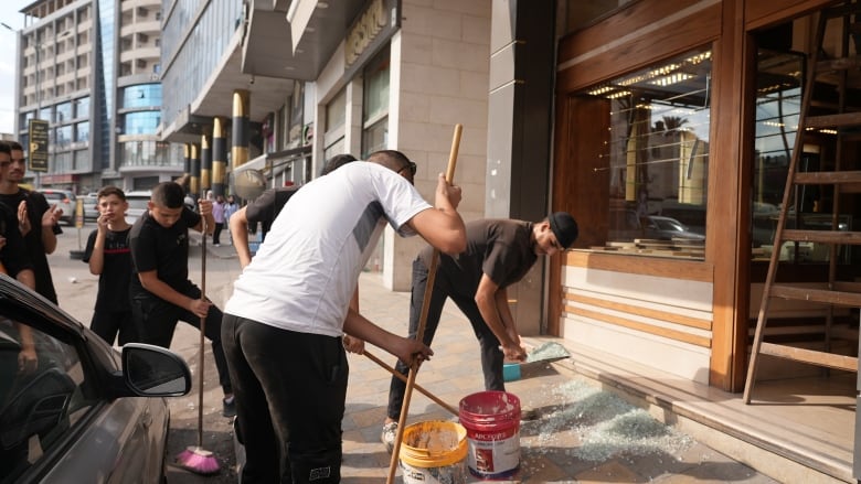 People use brooms and shovels to clear broken glass in front of a storefront.