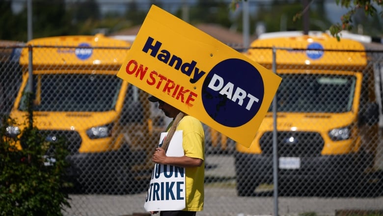 A person in a picket line with a couple of mini buses parked beyond a fence in the background.