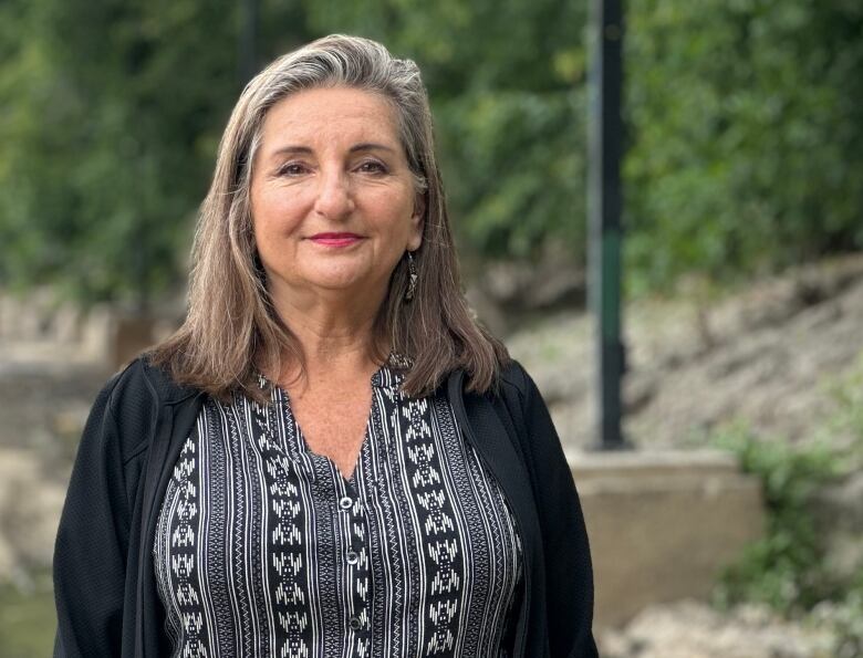 A white woman with shoulder length grey hair stands on the Assiniboine River trail.