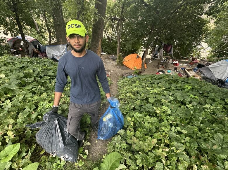 A man with a Downtown Community Safety partnership baseball cap stands in front of an encampment holding two garbage bags full of trash.