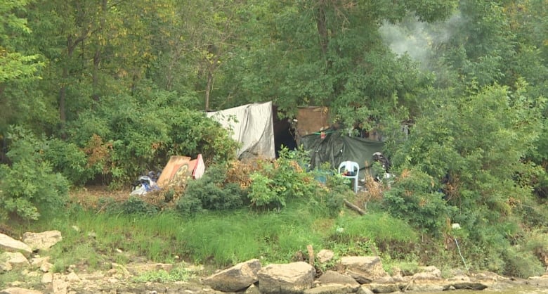 A few tents and a white plastic chair sit in the woods along the edge of the river, smoke from a campfire in the air.