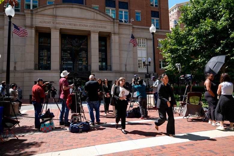 Lawyers and legal assistants leave a courthouse. News reporters and large cameras are gathered outside the stone building.