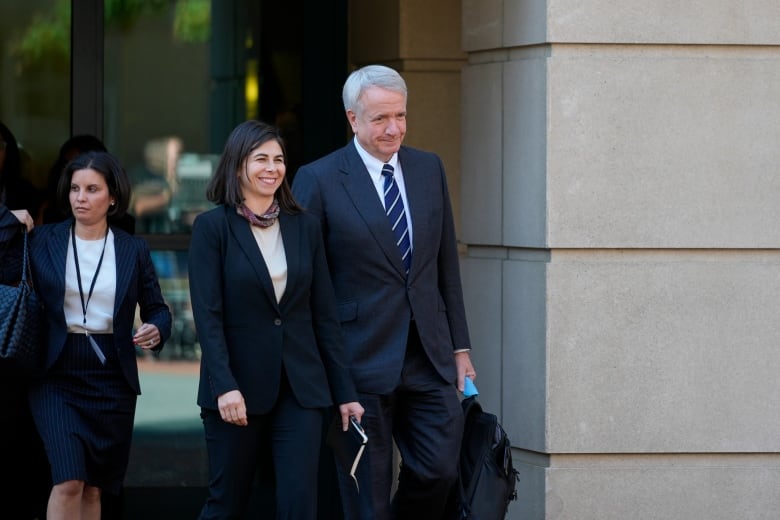 A man and a woman, both wearing suits, walk out of a courthouse. The man has white hair and the woman has straight, shoulder-length brown hair.