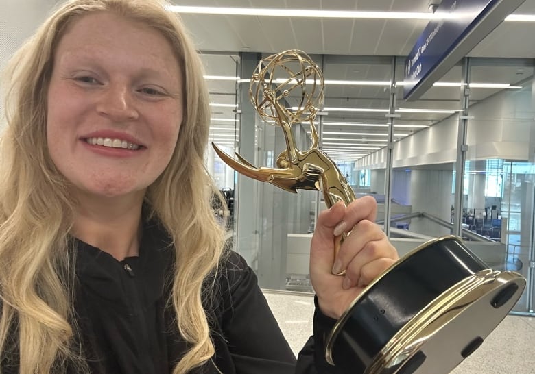 A smiling woman with blonde hair stands holding an Emmy trophy in an airport.