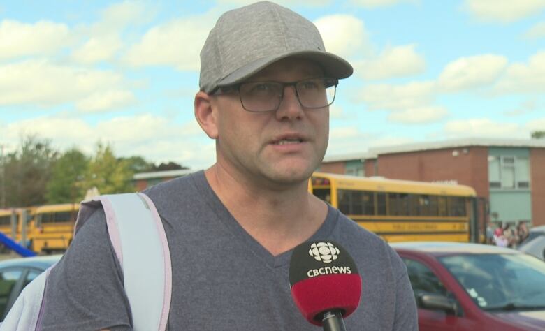 Man with ball cap and glasses talks into a TV news microphone as he stands in front of a yellow school bus and brick school building. 