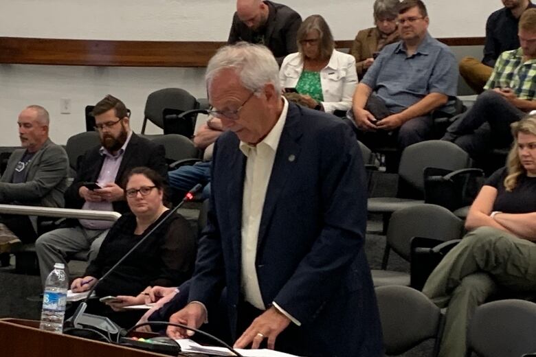 Man with grey hair wearing navy blue suit speaking at podium. 