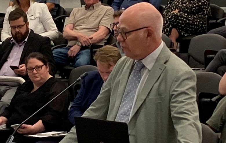 Bald man with glasses wearing light green blazer speaking at podium.