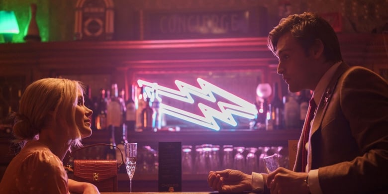 A young man and woman face each other, talking inside a neon-lit bar. 