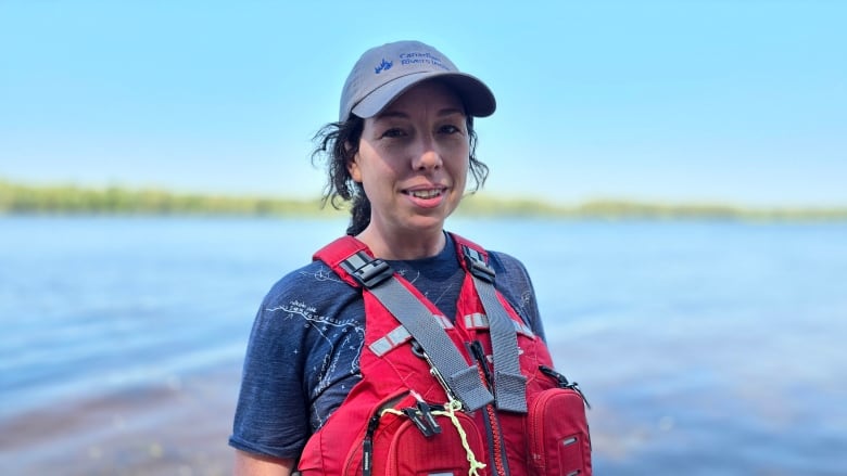 Woman in ballcap and lifejacket stands in front of a body of water. 