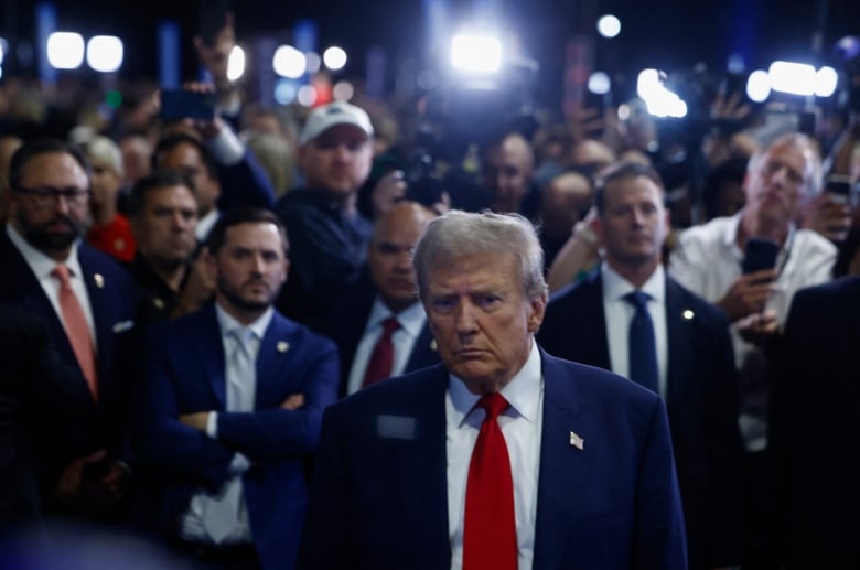 A man in a blue suit and red tie walks toward news reporters. A large crowd and bright lights are behind him.