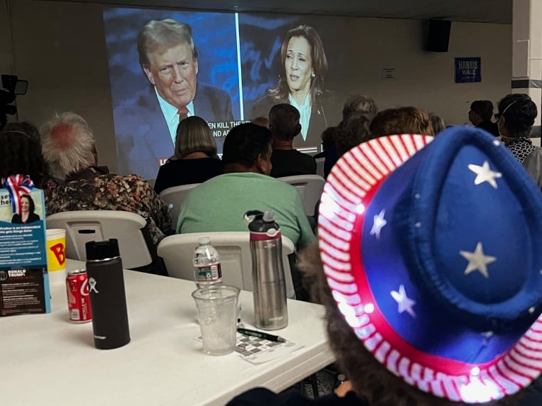 Woman in stars and stripes tophat watches screen showing Trump and Harris