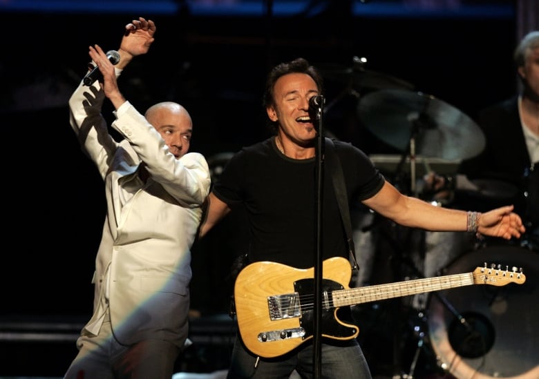 A man in a dark tshirt onstage with a guitar strapped around him spreads his arms as another man in white clothing dances, during a concert performance.