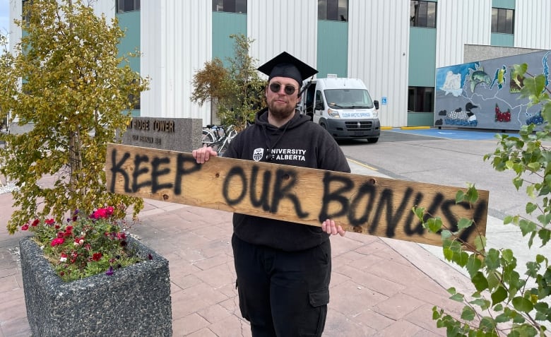 A man in a black hoodie and graduation cap holds a wooden sign spray-painted with the words 'KEEP OUR BONUS.'