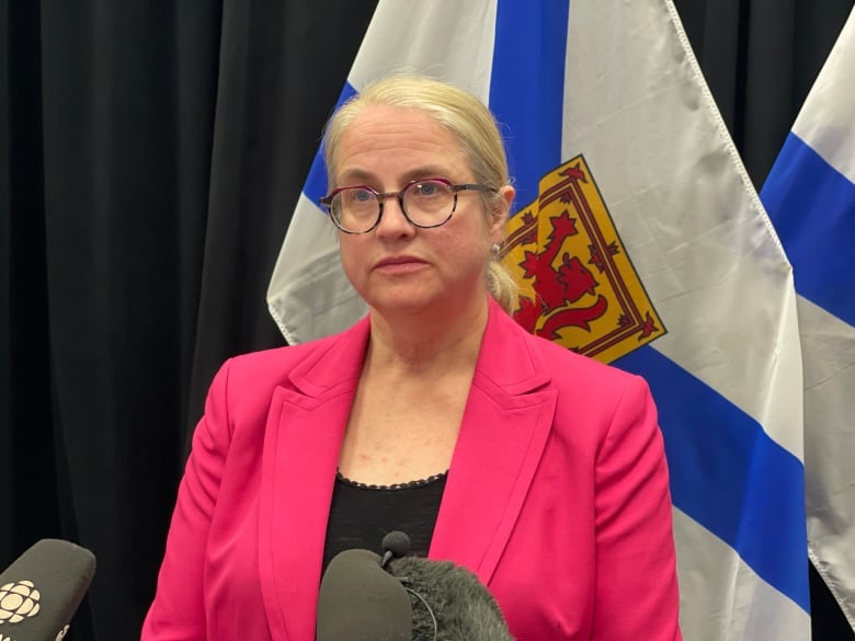 Woman in pink blazer and black top speaks stands in front of microphones.