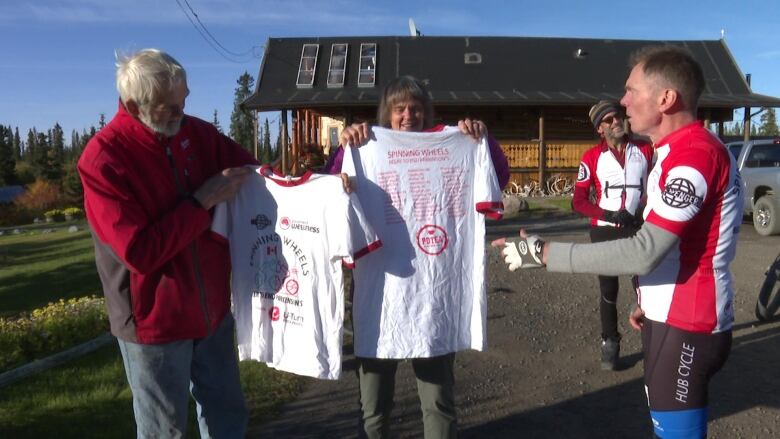 A few people stand outside, with 2 of them holding up t-shirts.