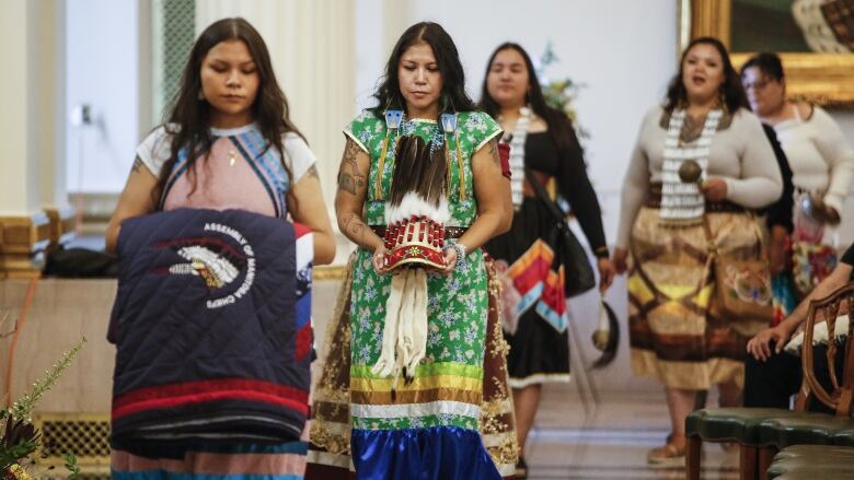 Women dressed in ribbon skirts carry items including a quilted blanket and a headdresss.