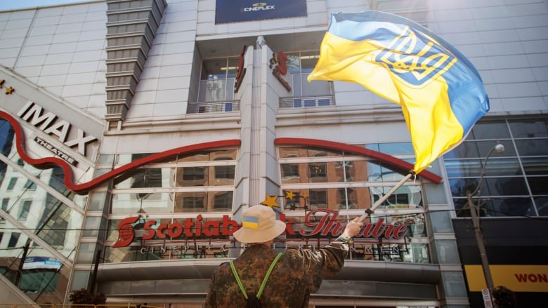 A man wearing military fatigues waves a red and blue flag in front of a movie theatre.