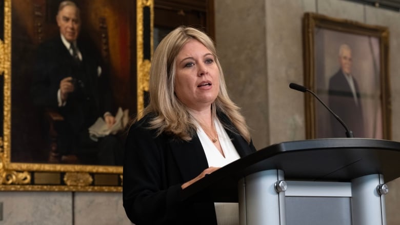 A woman in a black blazer and white shirt speaks in front of a podium.