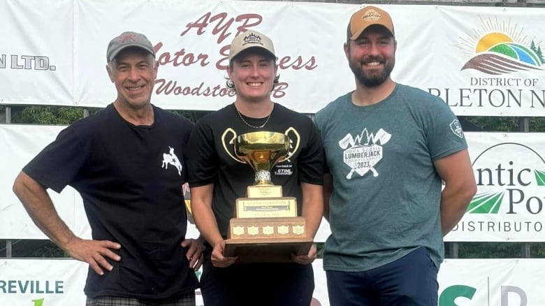 Three people smiling. The woman in the middle is holding a trophy.