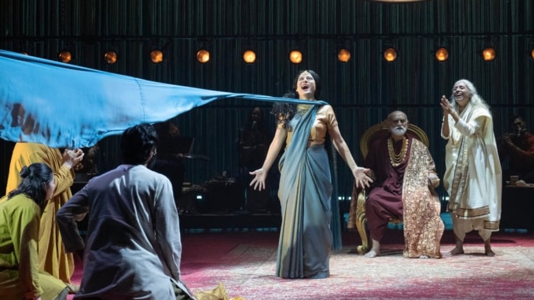 On a stage set beneath a chandelier, a group of actors  some standing, some sitting  look toward a woman at centre stage with a billowing dress.  