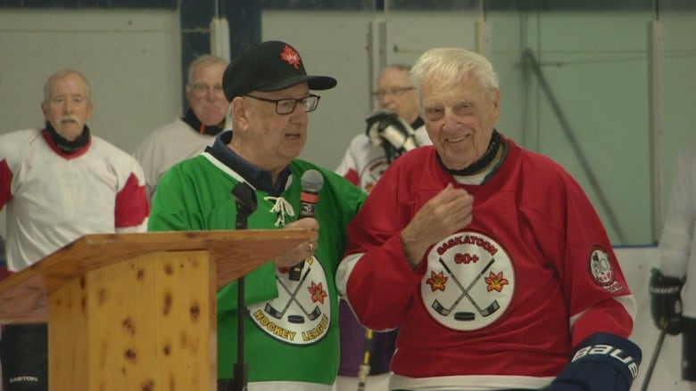 An older man in a green hockey jersey holds a microphone in front of a podium with his hand on the back of a smiling older man in a red hockey jersey.