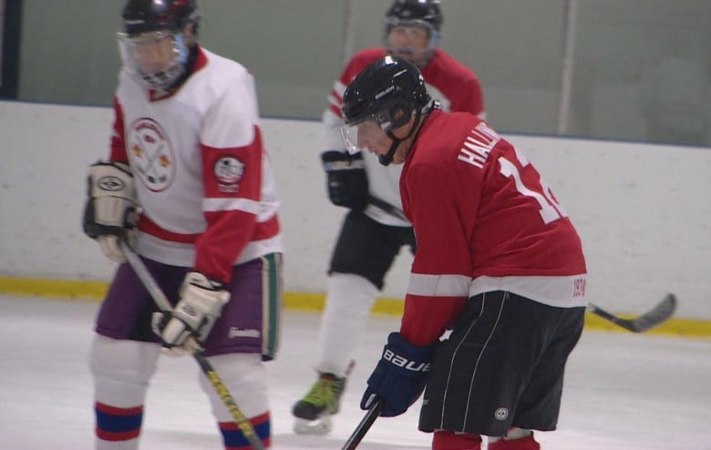 An older man wearing red, white, and black hockey gear waits for the puck to be dropped. Two opponents in white and red jerseys are in the background.