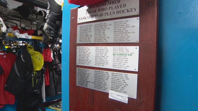 A plaque with the names of deceased hockey players on a blue wall in a hockey  dressing room. Hockey equipment hangs in stalls in the background.