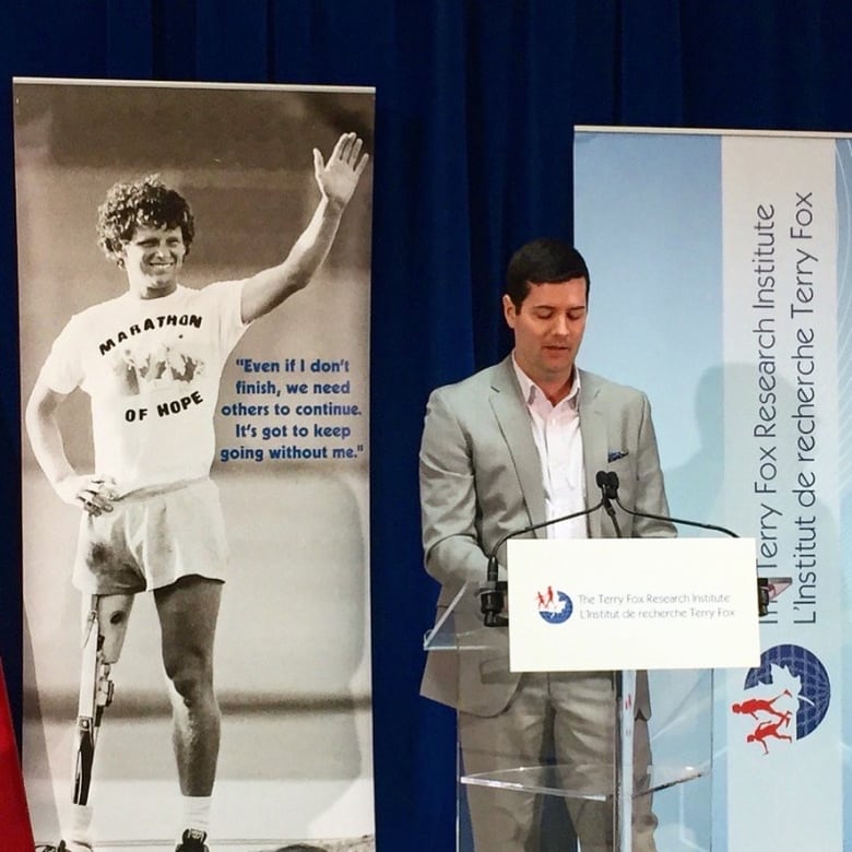 A man in a suit stands at a podium, reading a piece of paper and speaking into a microphone, with a large picture of Terry Fox on the stage behind him