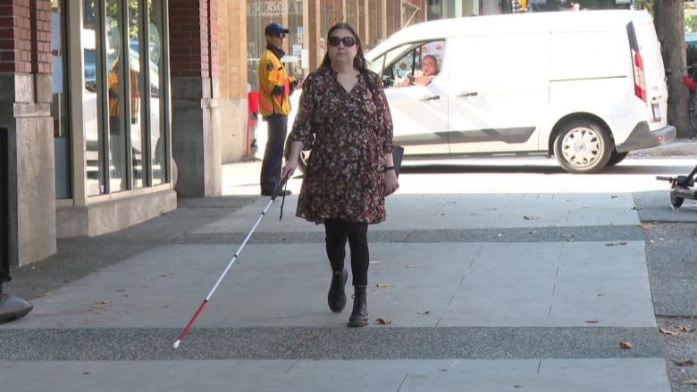 A woman with glasses and a white cane is pictured walking on a sidewalk. 