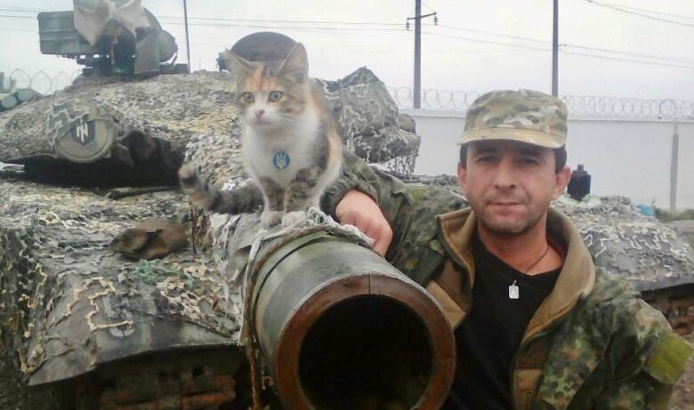 A man wearing a military cap and jacket stands beside a tank, as a cat sits on top.