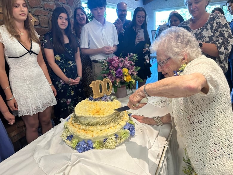 A woman cuts a cake with the number 100 on it, as people stand around and watch.