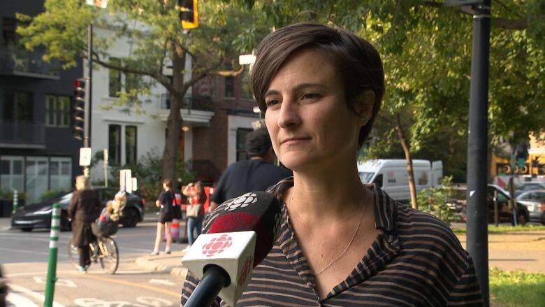 A portrait of a woman with short hair and a striped collared shirt during an interview outside, beside a bike lane. A CBC mic is held up to her face. 