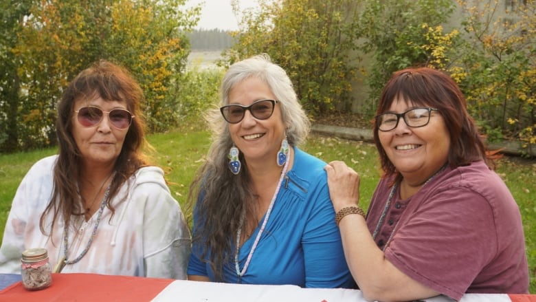 Three women sit at bench outside. 