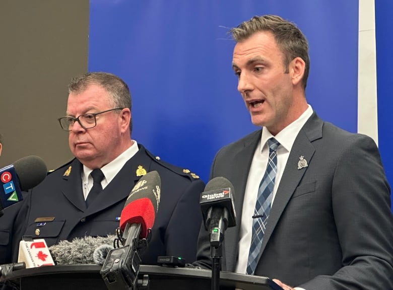 Two police officers stand in front of media microphones. One of the officers, wearing a grey suit with a blue argyle tie, is speaking. 