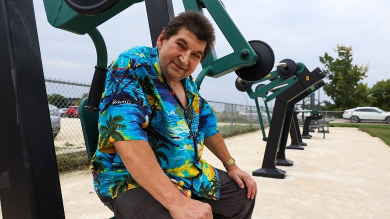 A man in a Hawaiian shirt sits on exercise equipment in a park.