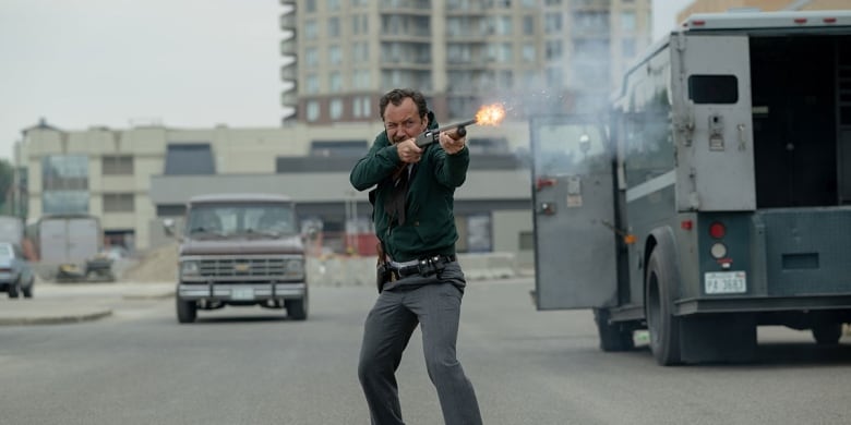 A man standing on a street fires a shotgun. There is an armoured truck parked behind him with both its front and rear doors open. 