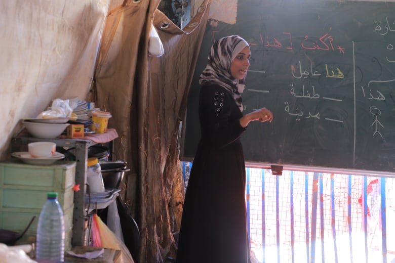 A woman in a black dress and grey headscarf stands at a chalkboard. 