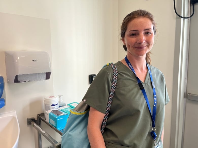 A physician poses in her green scrubs in a hospital room. Her personal effects bag strapped to her shoulder, and her hospital pass around her neck.