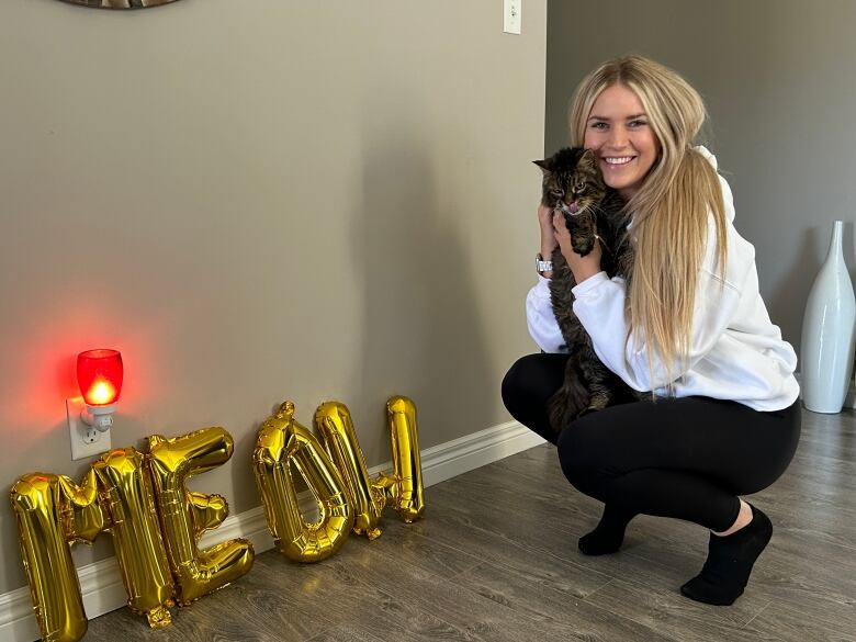 A blonde woman holds a brown cat. Balloons spelling out 