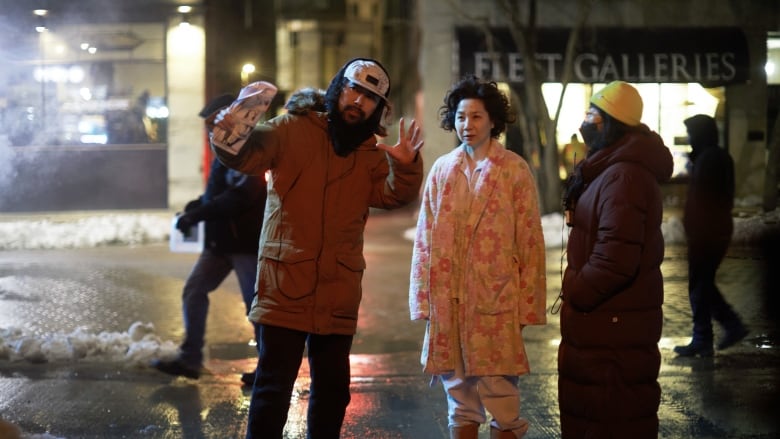 A man in a winter coat gestures as he stands on a city street at night next to a woman wearing a housecoat.