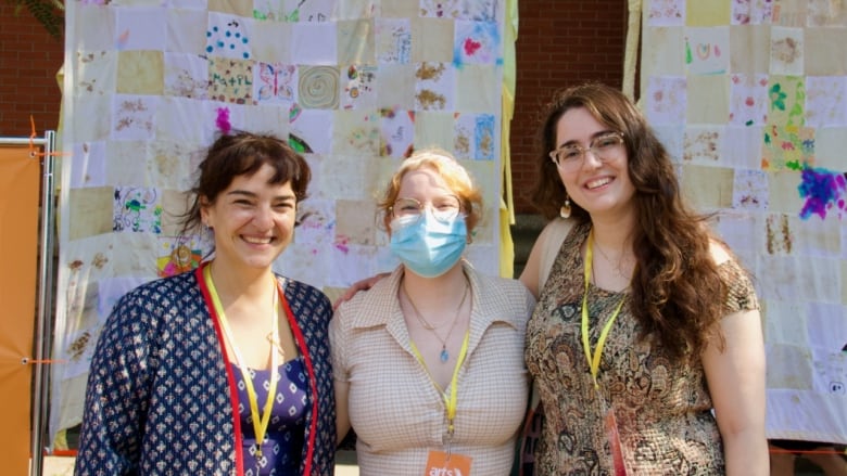 Three people with their arms around one another pose outdoors in front of a giant quilt. 