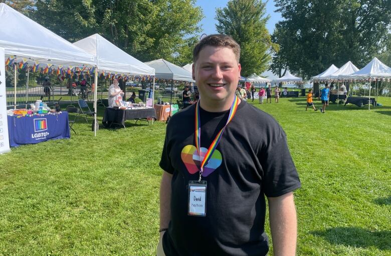 A man wearing a black shirt with two hearts. Patterns from different pride flags fill the hearts. He wears a rainbow lanyard. He's posing outside. Several white event tents are seen behind him. 