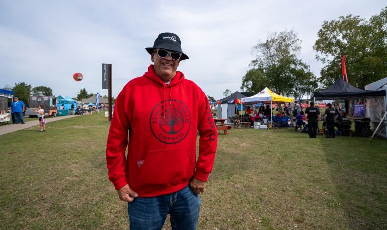 A person wearing a red hoodie and a black bucket hat stands outside, smiling.