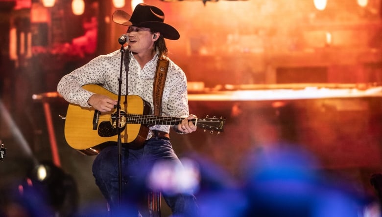 A man in a patterned white shirt and black cowboy hat plays a song while sitting on a stool.