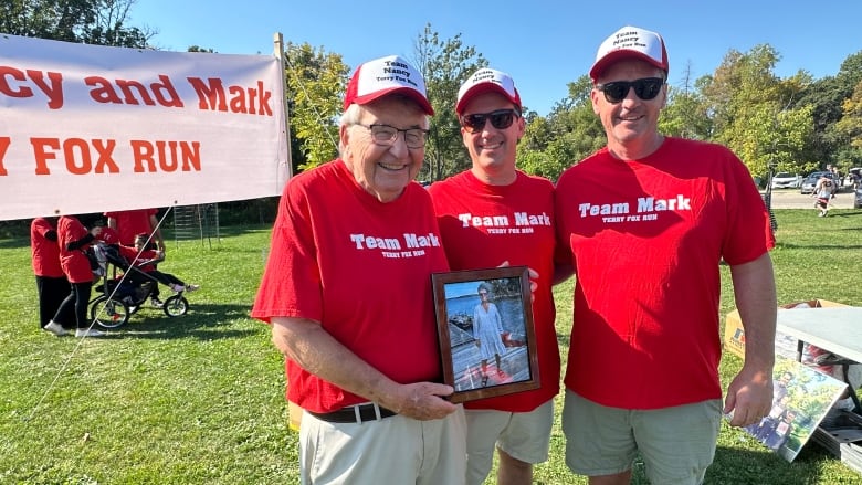 Three men wearing red t-shirts and hat pose for a photo outside. One of the men holds a picture frame of a woman.